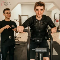 Mężczyzna na rowerze treningowym Assault Fitness w kombinezonie EMS, drugi mężczyzna z tabletem i stoperem obserwuje trening.