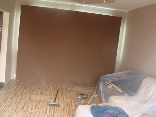 Pokój w trakcie remontu: świeżo pomalowana brązowa ściana, zabezpieczone meble folią, widoczna listwa przypodłogowa, parkiet i narzędzia malarskie.