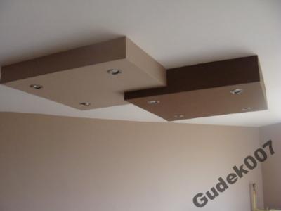 Dwukolorowy, kaskadowy sufit podwieszany z oświetleniem punktowym, beżowa ściana w tle.