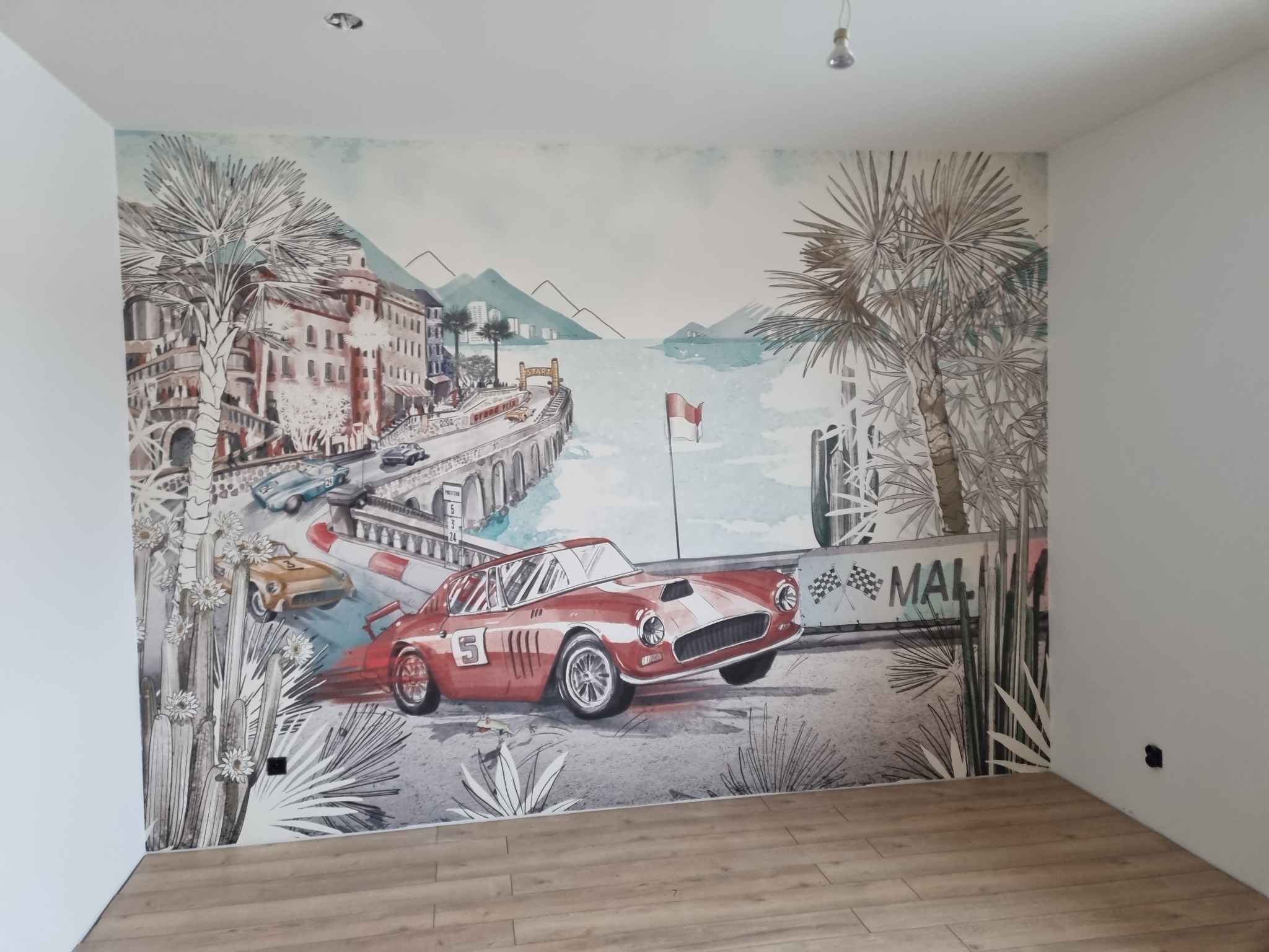Ściana w pokoju z fototapetą przedstawiającą scenę wyścigową z czerwonym samochodem sportowym, palmami i budynkami w stylu śródziemnomorskim, na podłodze panele imitujące drewno.
