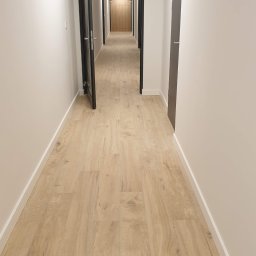 Usługi remontowe - Długi korytarz z panelami podłogowymi w odcieniach jasnego drewna, białe ściany z listwami przypodłogowymi i czarne ramy drzwi w perspektywie zbieżnej.