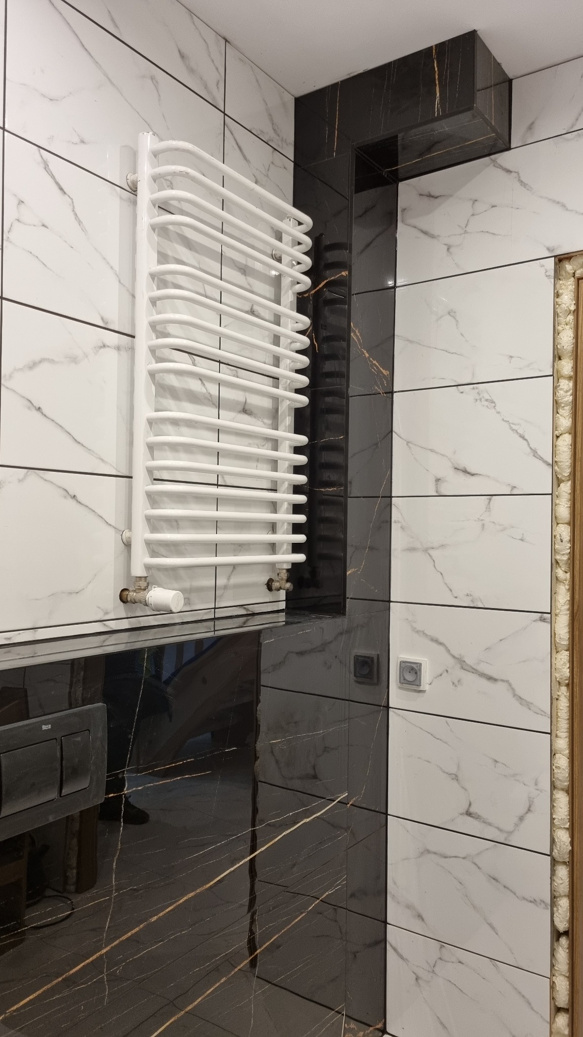 Biały grzejnik drabinkowy na tle ściany z płytek imitujących marmur w łazience z czarnymi akcentami.