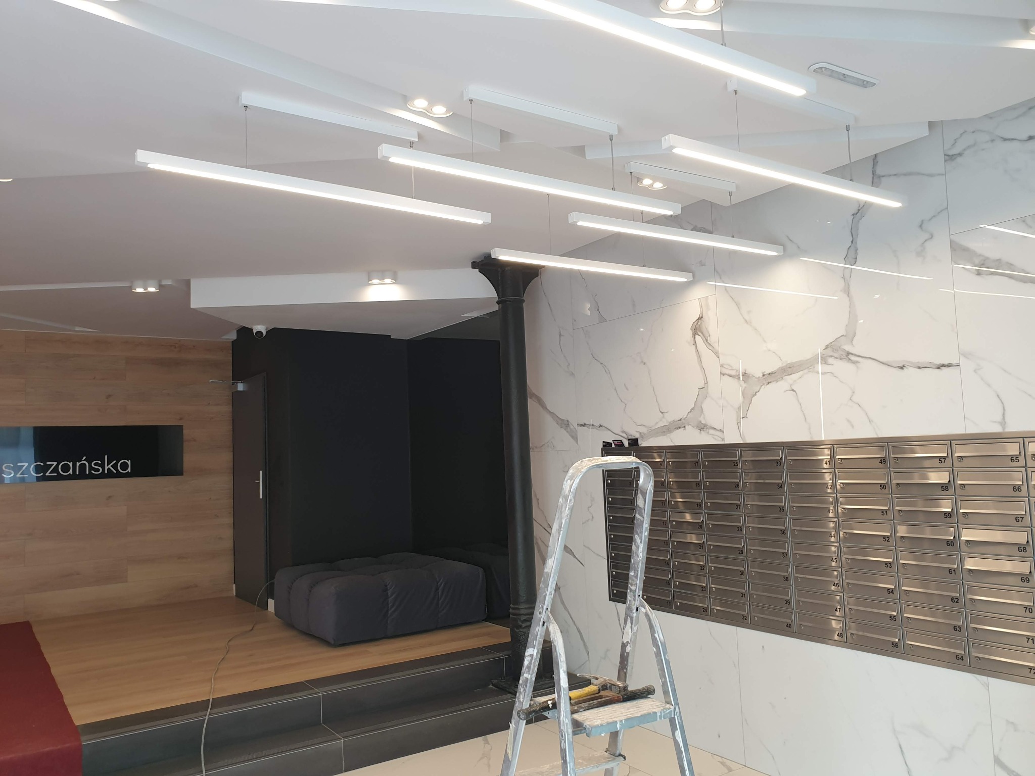 Nowoczesne oświetlenie sufitowe w holu budynku z marmurową ścianą, skrzynkami pocztowymi i drabiną malarską