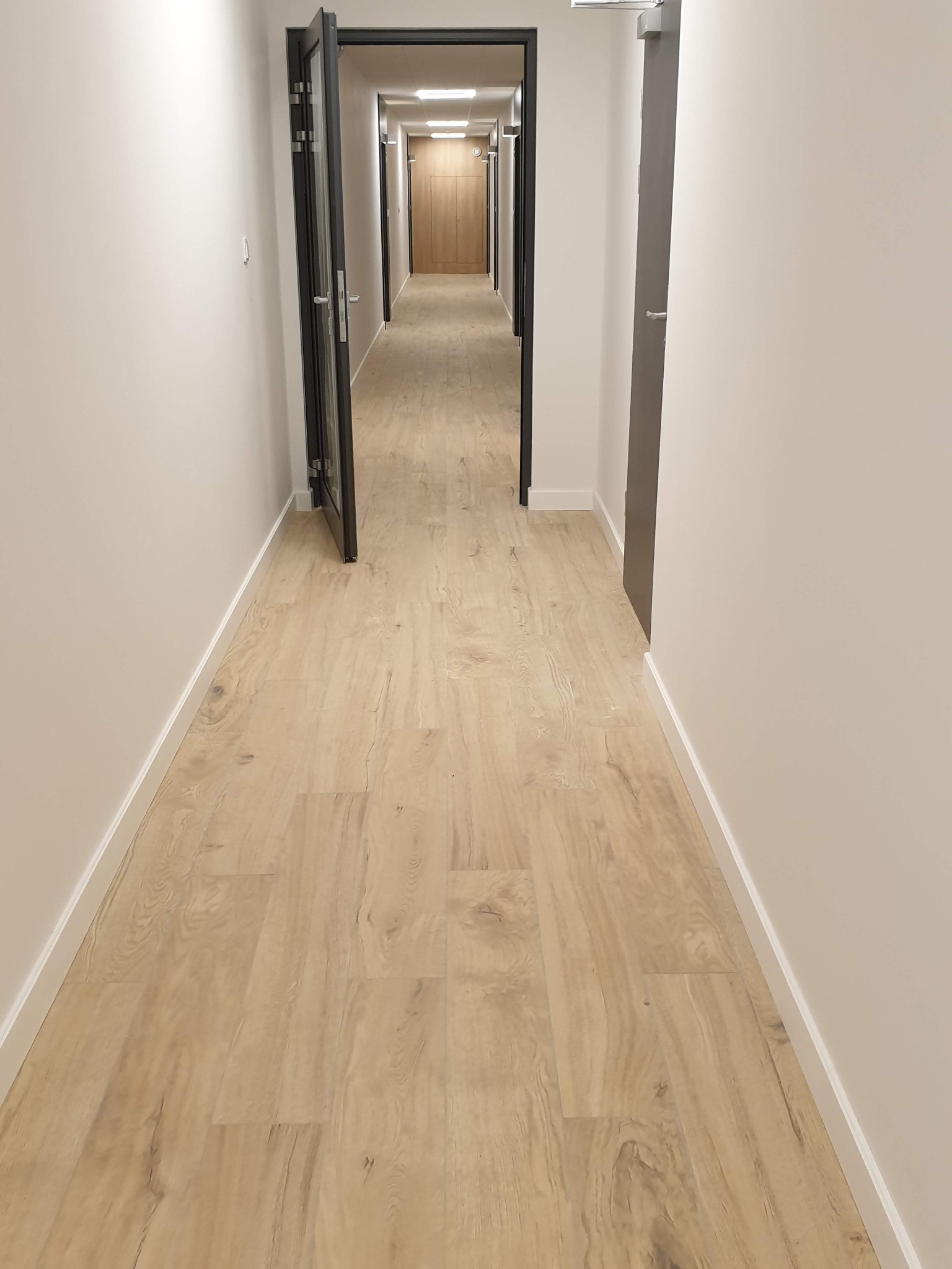 Długi korytarz z panelami podłogowymi w odcieniach jasnego drewna, białe ściany z listwami przypodłogowymi i czarne ramy drzwi w perspektywie zbieżnej.
