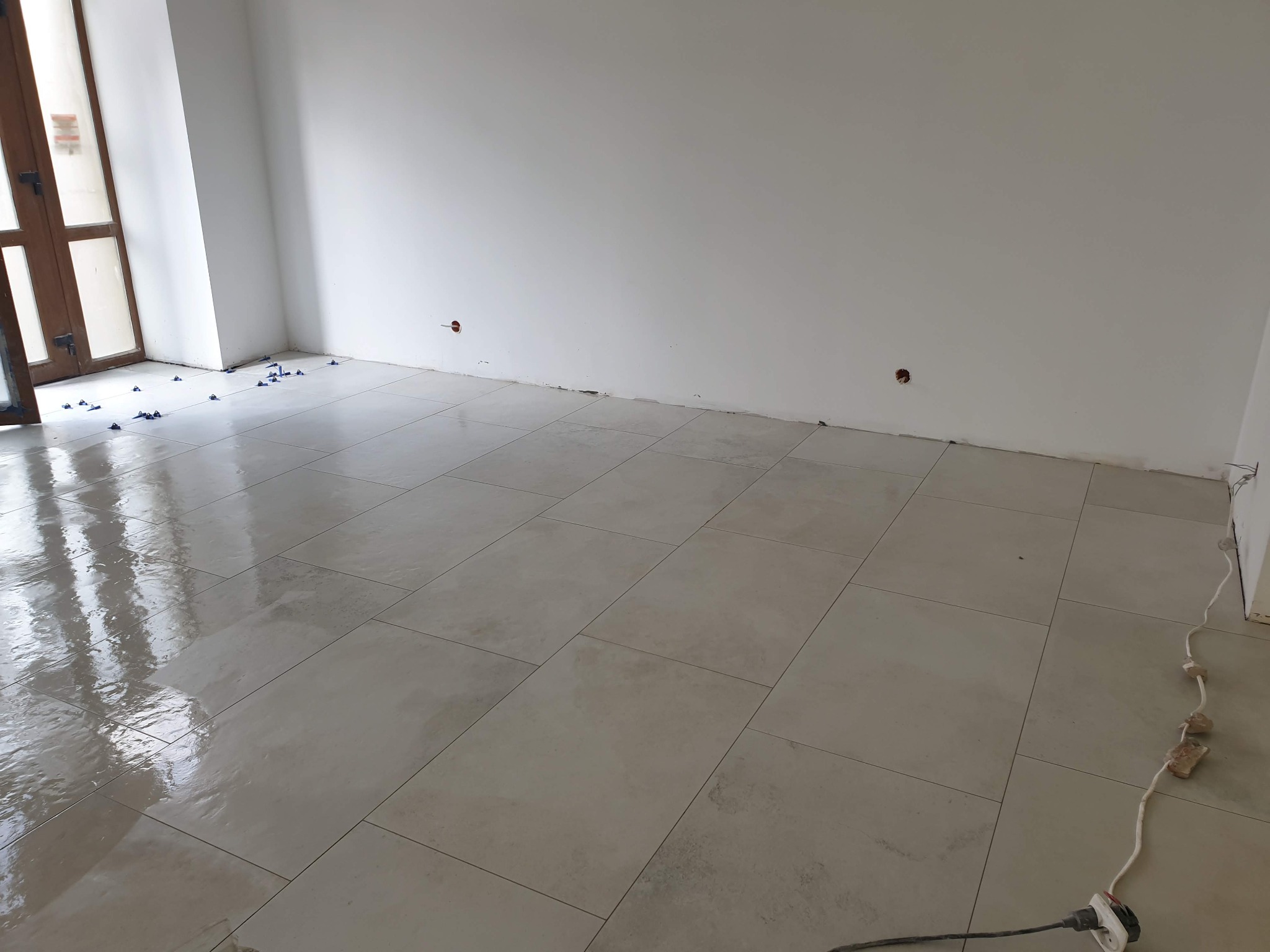 Świeżo położone, duże, beżowe płytki podłogowe w jasnym pomieszczeniu z widocznymi krzyżykami dystansowymi i kablem zasilającym. Na ścianach widoczne otwory po gniazdkach elektrycznych.