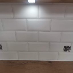 Teamlux - Białe, błyszczące płytki ułożone w cegiełkę nad drewnianym blatem we Wrocławiu, widoczne gniazdka elektryczne przed montażem osprzętu i punktowe oświetlenie podszafkowe.