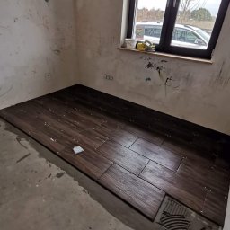 Teamlux - Układanie ciemnobrązowych płytek podłogowych imitujących drewno w trakcie prac wykończeniowych, widoczne krzyżyki dystansowe i resztki zaprawy, ściany z widocznymi śladami po rysunkach dziecięcych,...