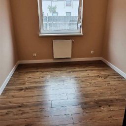 Teamlux - Pusty pokój z panelami podłogowymi w odcieniach brązu, beżowe ściany, biała listwa przypodłogowa i grzejnik pod oknem z widokiem na budynek mieszkalny.