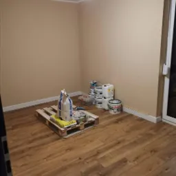 Pokój po malowaniu ścian na beżowo, z drewnianą podłogą, paletą z workami gładzi i farbami w wiaderkach przy ścianie.
