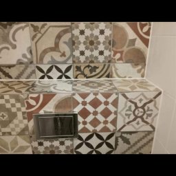 Teamlux - Ściana i obudowa stelaża WC wyłożone mozaiką z płytek ceramicznych o różnorodnych wzorach w odcieniach beżu, brązu i szarości, z widocznym chromowanym przyciskiem spłukującym.