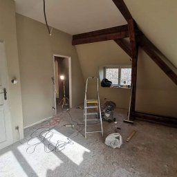 Teamlux - Pokój na poddaszu w trakcie remontu: świeżo pomalowane ściany w odcieniach beżu, widoczne drewniane belki stropowe, rozłożona drabina aluminiowa, kable elektryczne na podłodze i lampa budowlana w tle.