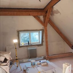 Teamlux - Poddasze w trakcie prac wykończeniowych, widoczne drewniane belki konstrukcyjne, ściany po nałożeniu gładzi, okno z parapetem, grzejnik, lampa budowlana na trójnogu oraz materiały i narzędzia...