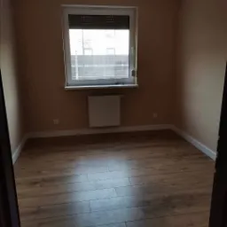 Pusty pokój po remoncie z panelami podłogowymi w odcieniach brązu, świeżo pomalowanymi ścianami w kolorze beżowym, białymi listwami przypodłogowymi i grzejnikiem pod oknem z roletami.
