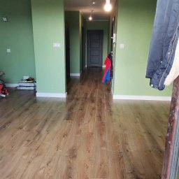 Teamlux - Korytarz po remoncie z zielonymi ścianami, drewnopodobną podłogą i widocznymi elementami osobistymi, takimi jak ubrania i zabawki, sugerujący zamieszkanie.