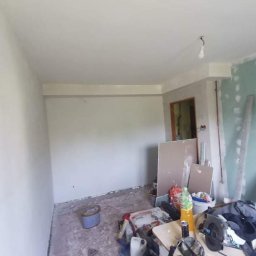 Teamlux - Pokój w trakcie remontu: świeżo pomalowane ściany na biało, widoczne narzędzia malarskie, płyty gipsowe oparte o ścianę, bałagan na podłodze.