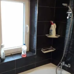 Biała wanna w łazience z czarnymi płytkami imitującymi drewno, półki z detergentami i butelki na parapecie okna.