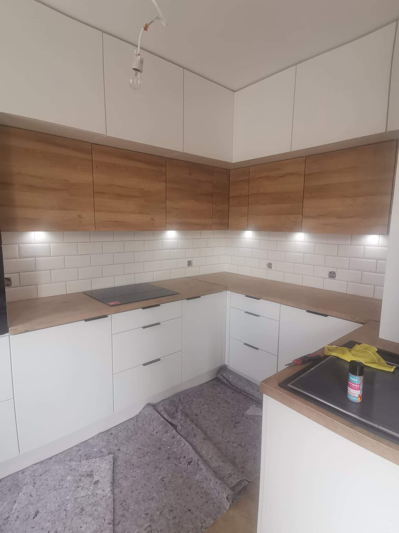 Montaż kuchni: białe szafki dolne z ciemnymi uchwytami, górne szafki w odcieniu drewna, białe płytki na ścianie, płyta indukcyjna, pod szafkami zamontowane oświetlenie LED, podłoga zabezpieczona...