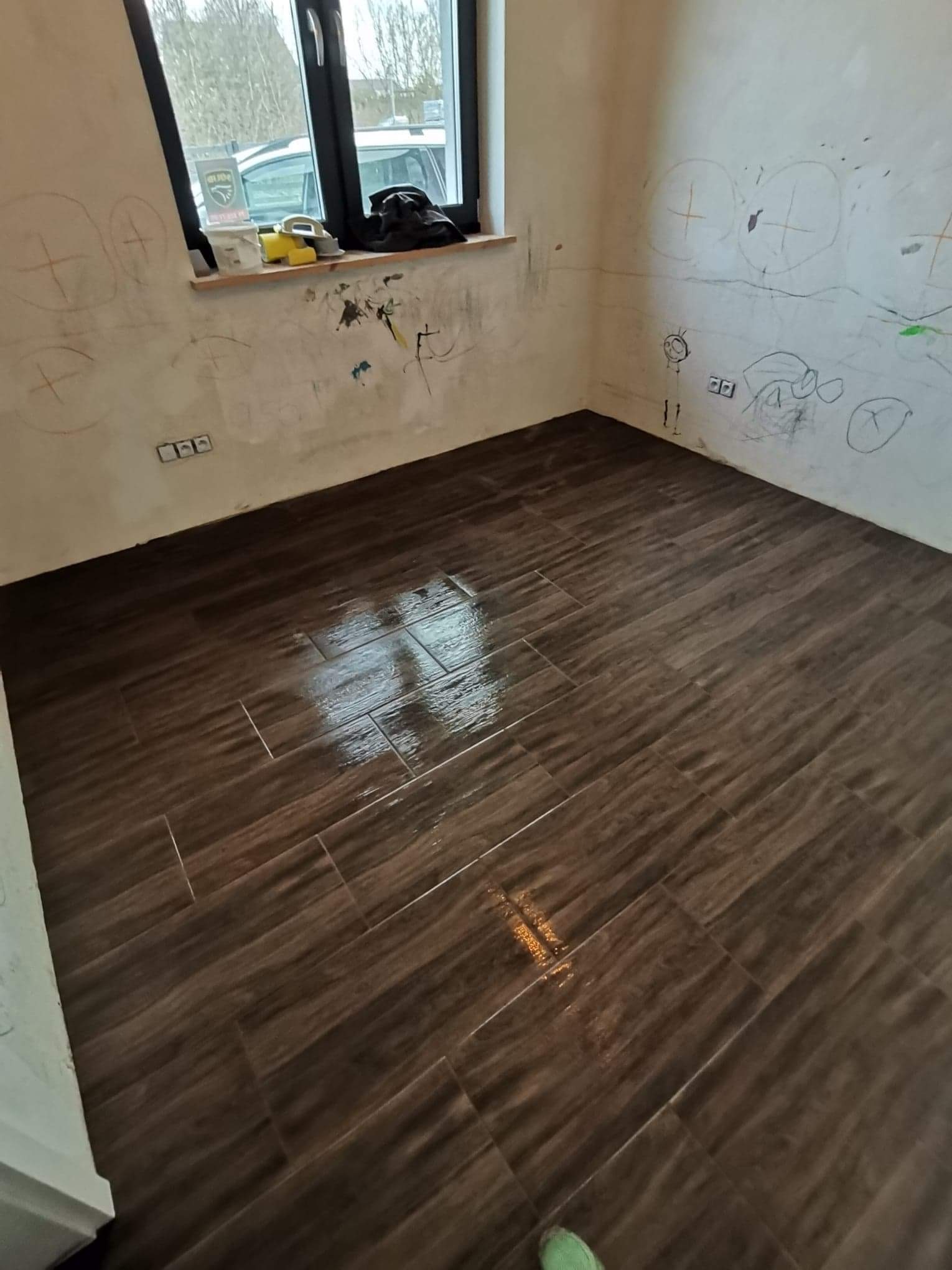Ułożona, brązowa podłoga z płytek imitujących drewno w pokoju z pomalowanymi przez dzieci ścianami. Widoczne ślady po rysunkach na ścianach i akcesoria na parapecie okiennym.
