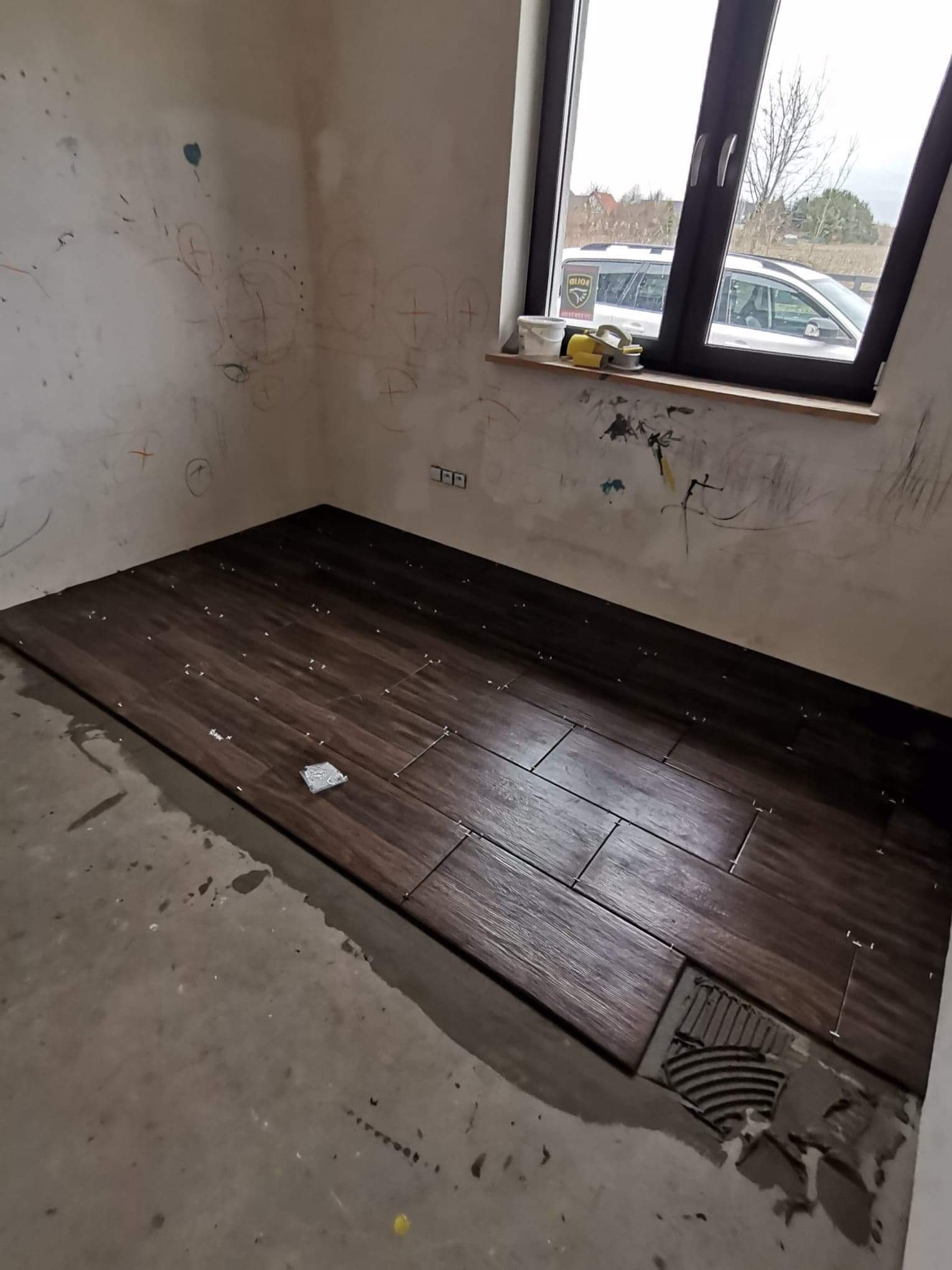 Układanie ciemnobrązowych płytek podłogowych imitujących drewno w trakcie prac wykończeniowych, widoczne krzyżyki dystansowe i resztki zaprawy, ściany z widocznymi śladami po rysunkach dziecięcych,...