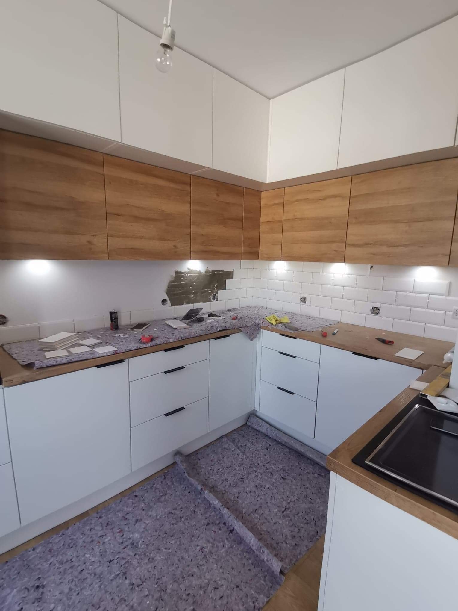 Montaż białej glazury w kuchni z drewnianymi szafkami górnymi, widoczne płytki na blacie i zabezpieczenie podłogi folią ochronną.