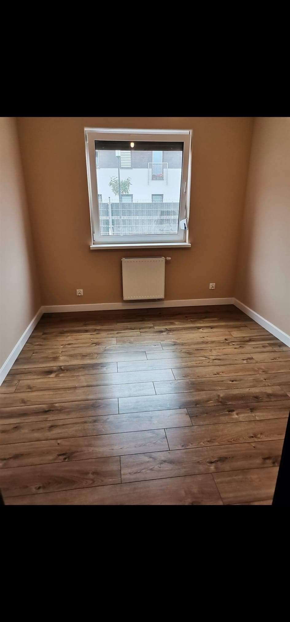 Pusty pokój z panelami podłogowymi w odcieniach brązu, beżowe ściany, biała listwa przypodłogowa i grzejnik pod oknem z widokiem na budynek mieszkalny.