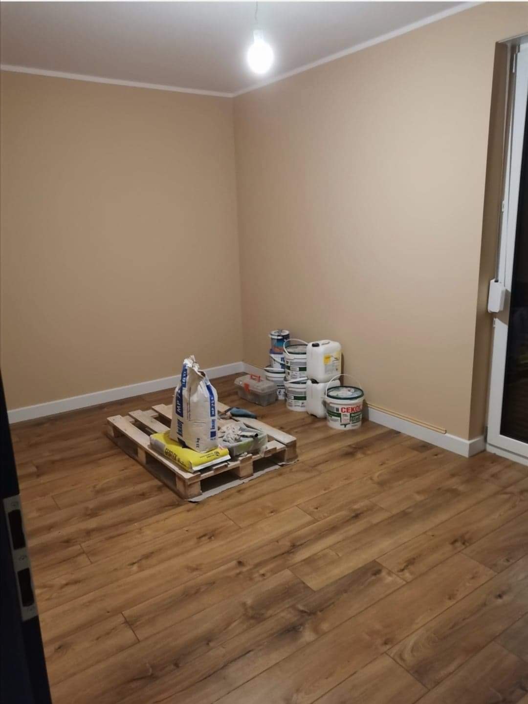 Pokój po malowaniu ścian na beżowo, z drewnianą podłogą, paletą z workami gładzi i farbami w wiaderkach przy ścianie.