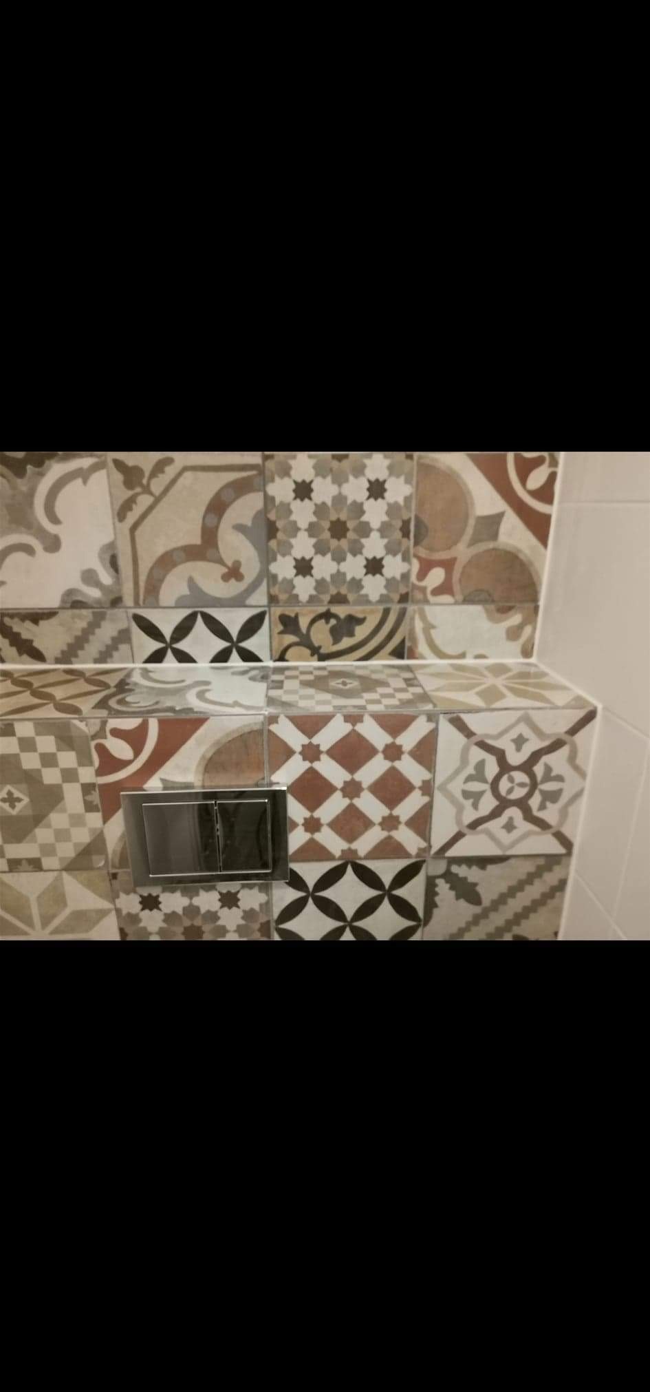 Ściana i obudowa stelaża WC wyłożone mozaiką z płytek ceramicznych o różnorodnych wzorach w odcieniach beżu, brązu i szarości, z widocznym chromowanym przyciskiem spłukującym.