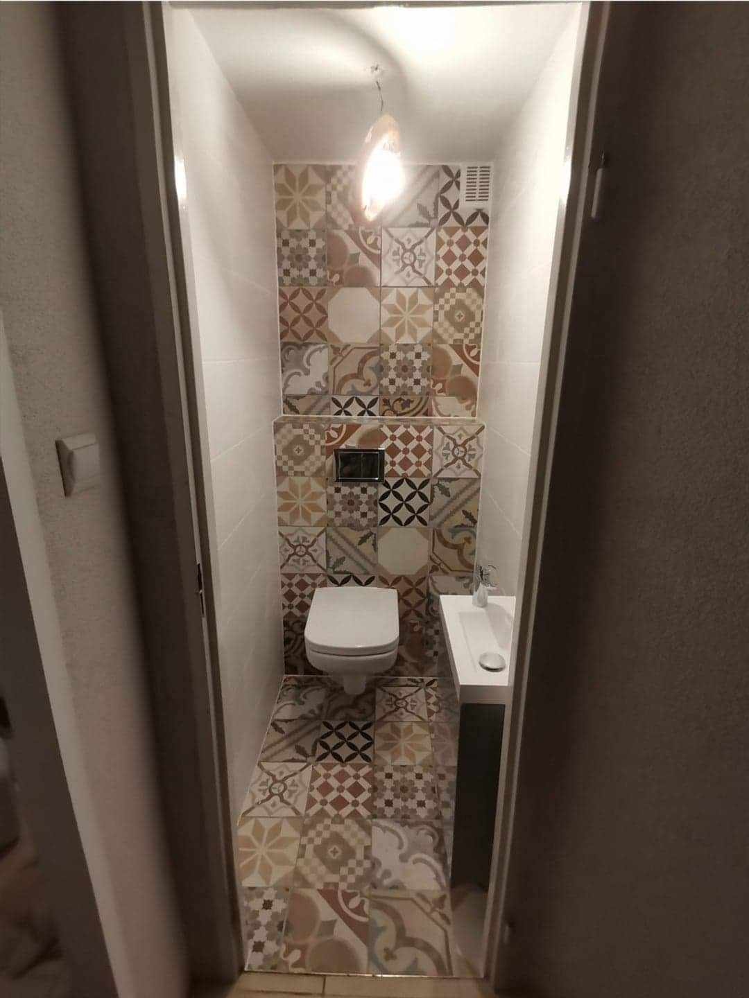 Mała łazienka z toaletą podwieszaną, umywalką nablatową i mozaikową okładziną ścian i podłogi w stylu patchwork, oświetlona pojedynczą żarówką.