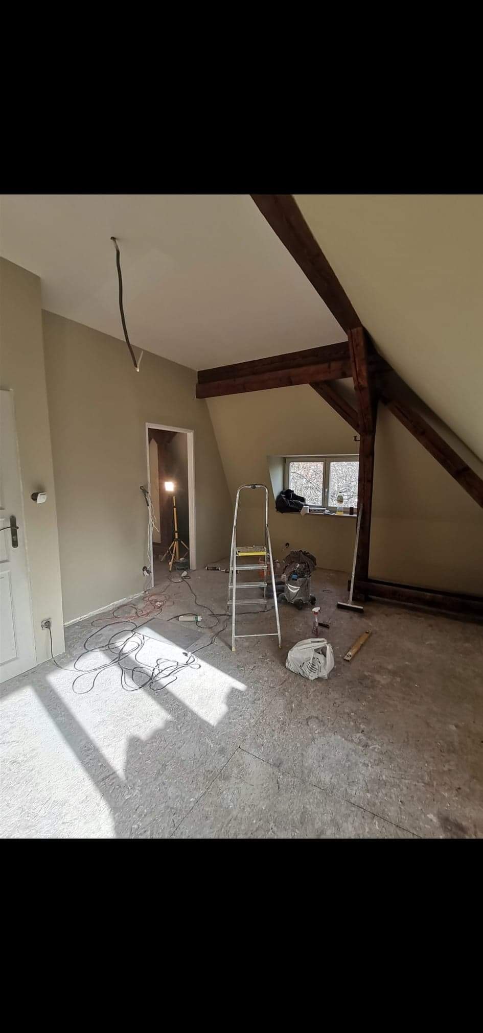 Pokój na poddaszu w trakcie remontu: świeżo pomalowane ściany w odcieniach beżu, widoczne drewniane belki stropowe, rozłożona drabina aluminiowa, kable elektryczne na podłodze i lampa budowlana w tle.