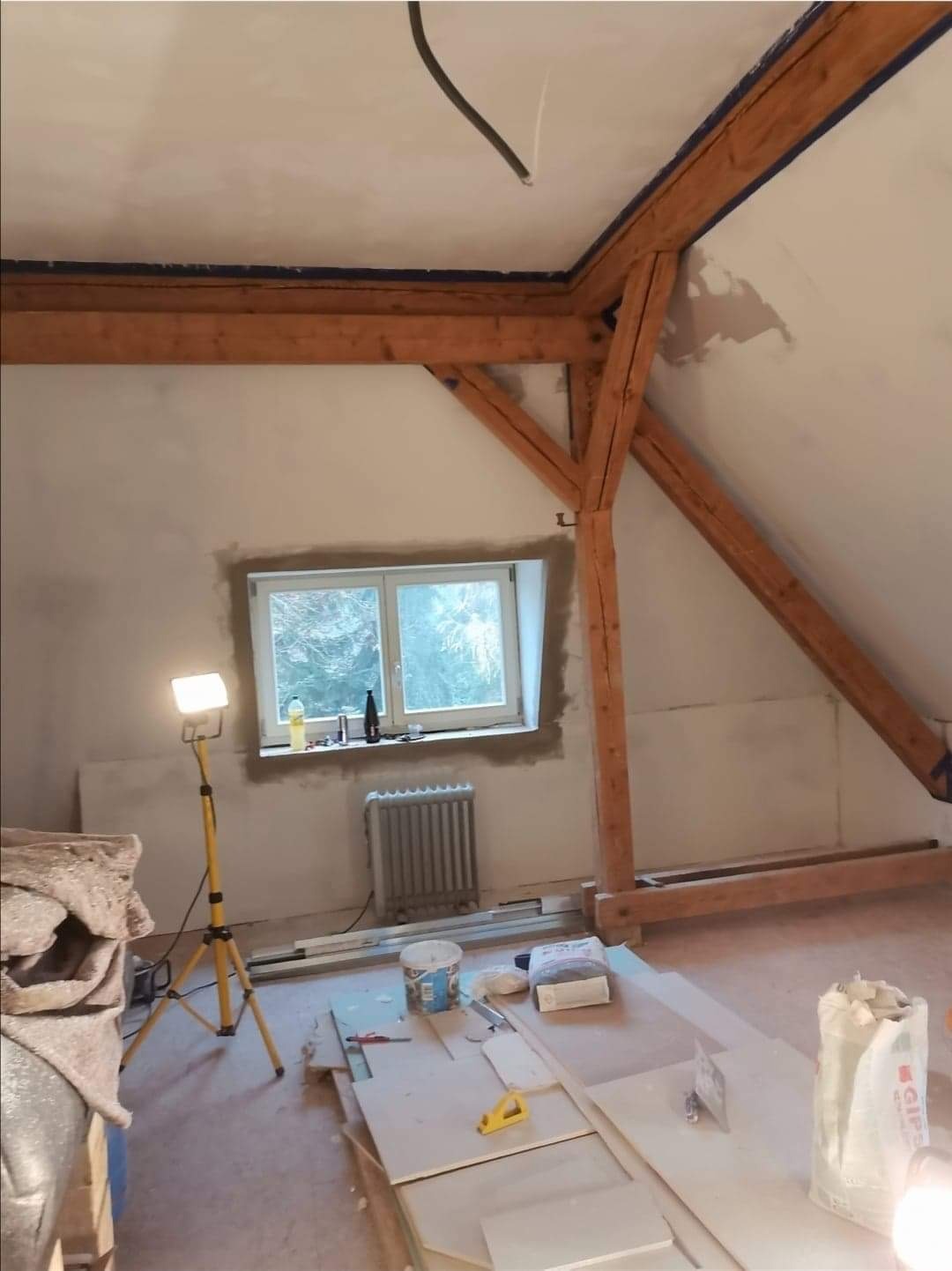 Poddasze w trakcie prac wykończeniowych, widoczne drewniane belki konstrukcyjne, ściany po nałożeniu gładzi, okno z parapetem, grzejnik, lampa budowlana na trójnogu oraz materiały i narzędzia...