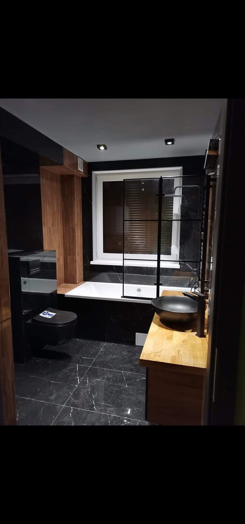 Nowoczesna łazienka z czarnymi marmurowymi płytkami, drewnianymi akcentami i czarną armaturą, ukazująca umywalkę nablatową, wiszącą toaletę i wannę z parawanem.