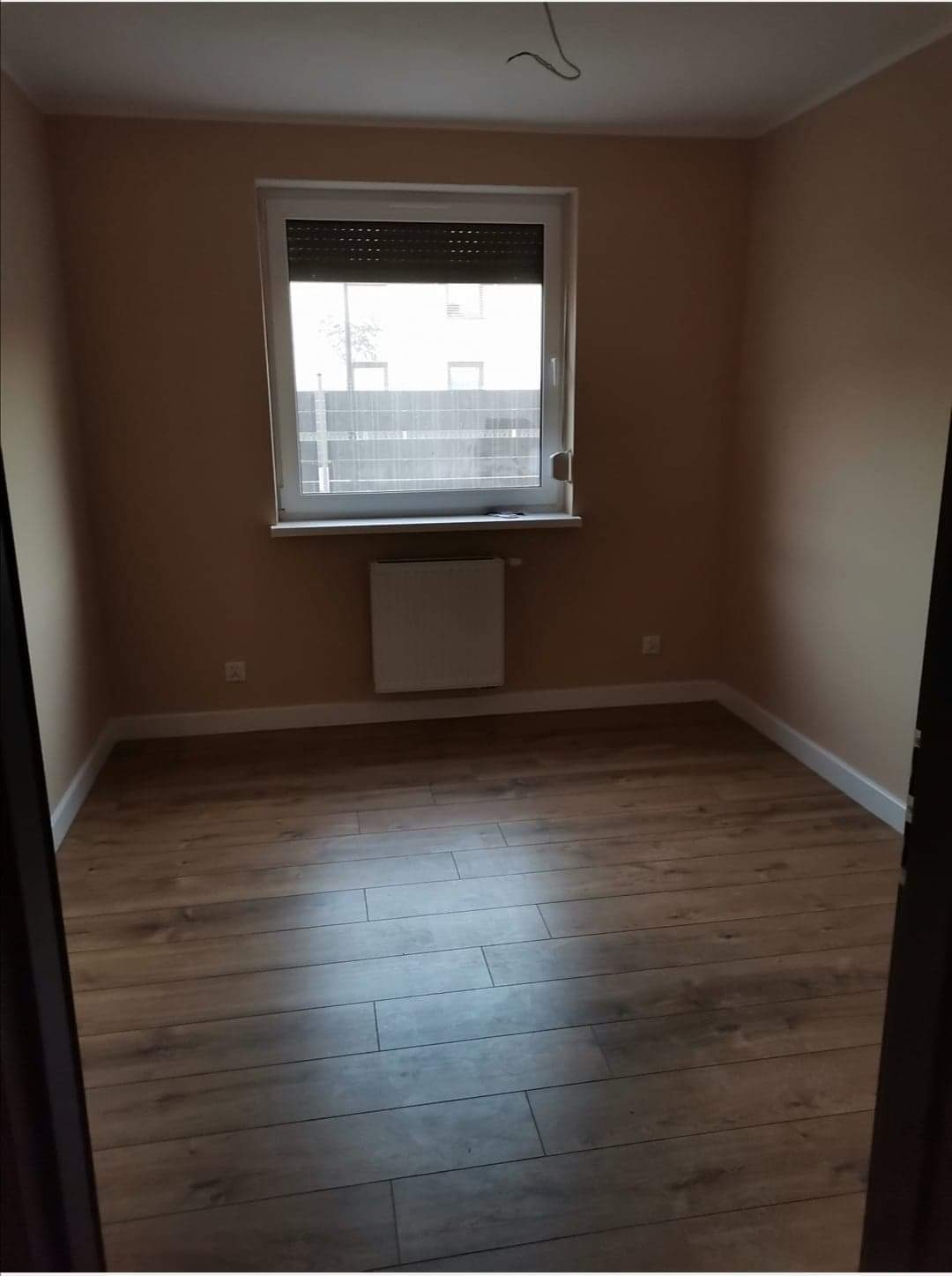 Pusty pokój po remoncie z panelami podłogowymi w odcieniach brązu, świeżo pomalowanymi ścianami w kolorze beżowym, białymi listwami przypodłogowymi i grzejnikiem pod oknem z roletami.