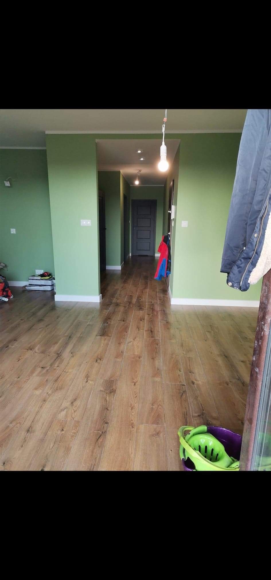 Korytarz po remoncie z zielonymi ścianami, drewnopodobną podłogą i widocznymi elementami osobistymi, takimi jak ubrania i zabawki, sugerujący zamieszkanie.