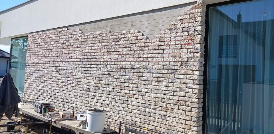 Kładzenie jasnej okładziny z imitacji kamienia na elewacji budynku, widoczne fragmenty niezakończonej pracy i narzędzia na rusztowaniu.