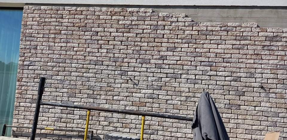 Ściana zewnętrzna budynku w trakcie wykańczania z częściowo ułożoną dekoracyjną okładziną imitującą cegłę w odcieniach szarości i beżu, widoczne rusztowanie i fragment niezakończonej powierzchni.