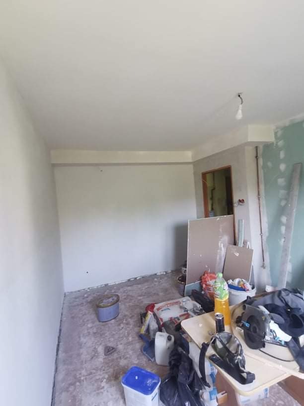 Pokój w trakcie remontu: świeżo pomalowane ściany na biało, widoczne narzędzia malarskie, płyty gipsowe oparte o ścianę, bałagan na podłodze.