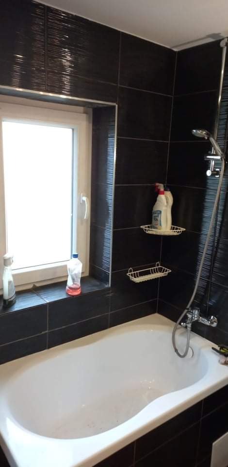 Biała wanna w łazience z czarnymi płytkami imitującymi drewno, półki z detergentami i butelki na parapecie okna.