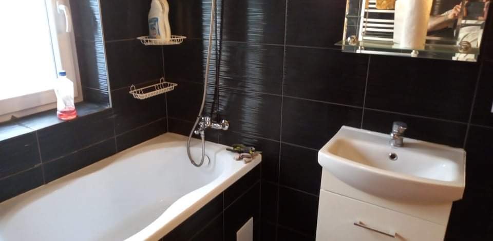 Ciemna łazienka po renowacji z czarnymi błyszczącymi płytkami, białą wanną z chromowaną armaturą, umywalką na szafce i półkami na ścianie.