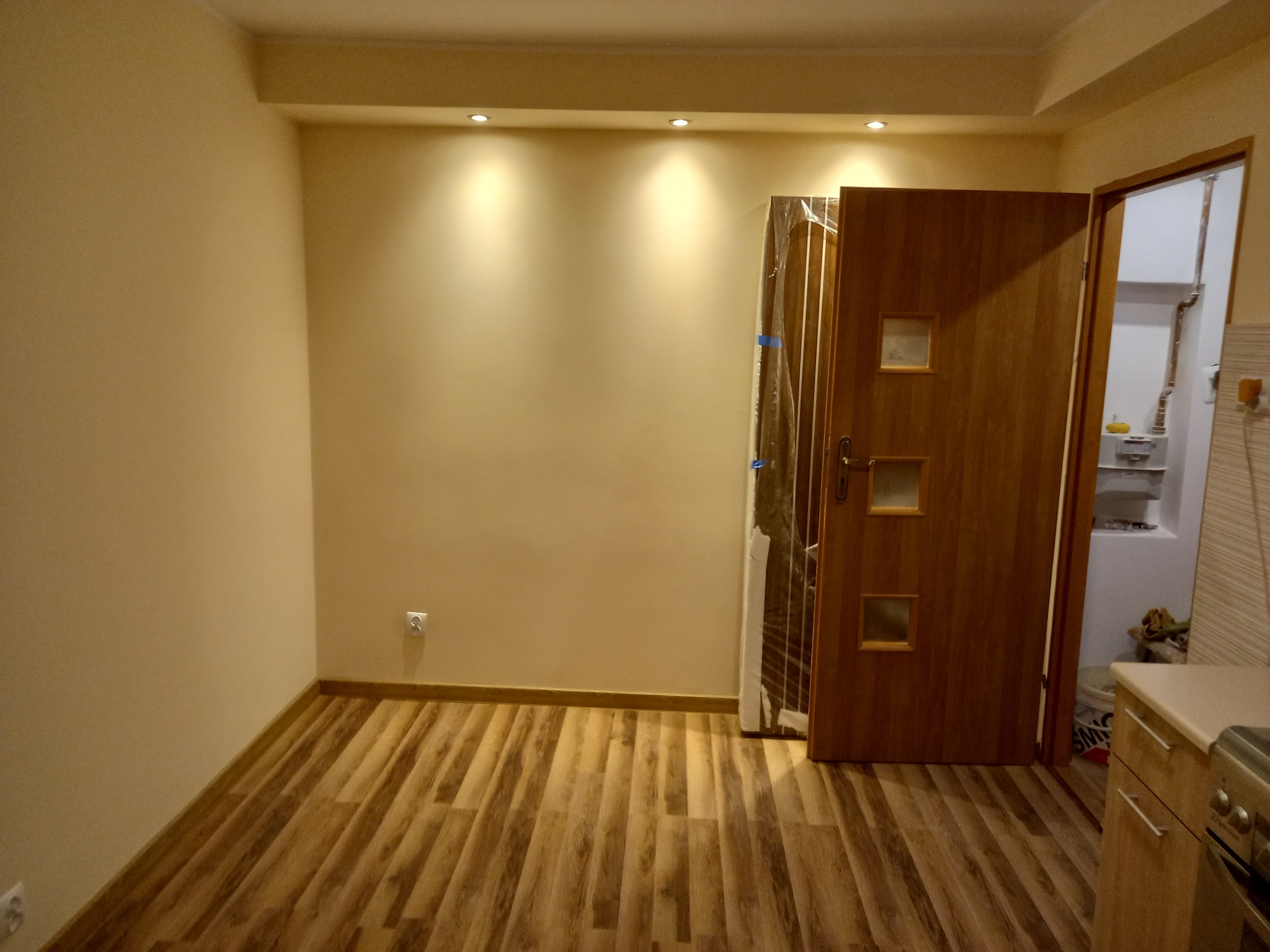 Pusty pokój z nową podłogą panelową w odcieniach brązu, ściany pomalowane na beżowo, widoczne punkty świetlne w suficie, częściowo zasłonięte drzwi z trzema przeszkleniami, zabezpieczone folią...