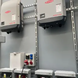 Dwa falowniki Fronius SYMO zamontowane na szynach, poniżej rozdzielnice AC i DC z ostrzeżeniami o napięciu, instalacja elektryczna w szarej ścianie.