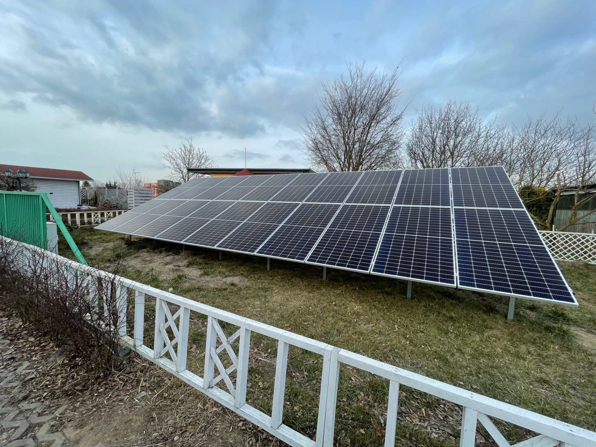 Rząd paneli słonecznych na trawiastym terenie, widoczny zza białego, drewnianego płotu z ozdobnymi krzyżami, z budynkami w tle i pochmurnym niebem.