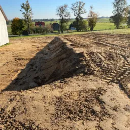 Przygotowany teren pod budowę domu, z widocznymi śladami opon ciężkiego sprzętu i świeżo wykopanym głębokim rowem, otoczony polami i drzewami w słoneczny dzień.