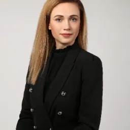 Portret kobiety w czarnym żakiecie z metalowymi guzikami i czarnej bluzce z falbanką pod szyją, jasne włosy opadają na ramiona, neutralne tło.