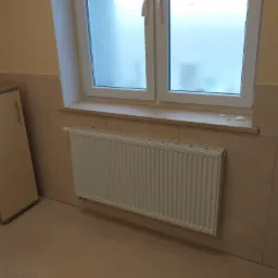 Ściana wyłożona płytkami w odcieniach beżu, biały grzejnik pod oknem z mleczną szybą, obok szafka z jasnego drewna.