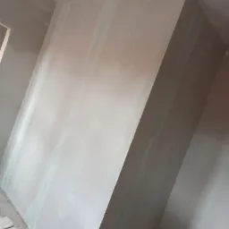 Skośna ściana z płyt kartonowo-gipsowych, z widocznymi łączeniami i śladami szpachlowania, przygotowana do dalszych prac wykończeniowych, z widocznym fragmentem podłogi i palety z cegłami w lewym...