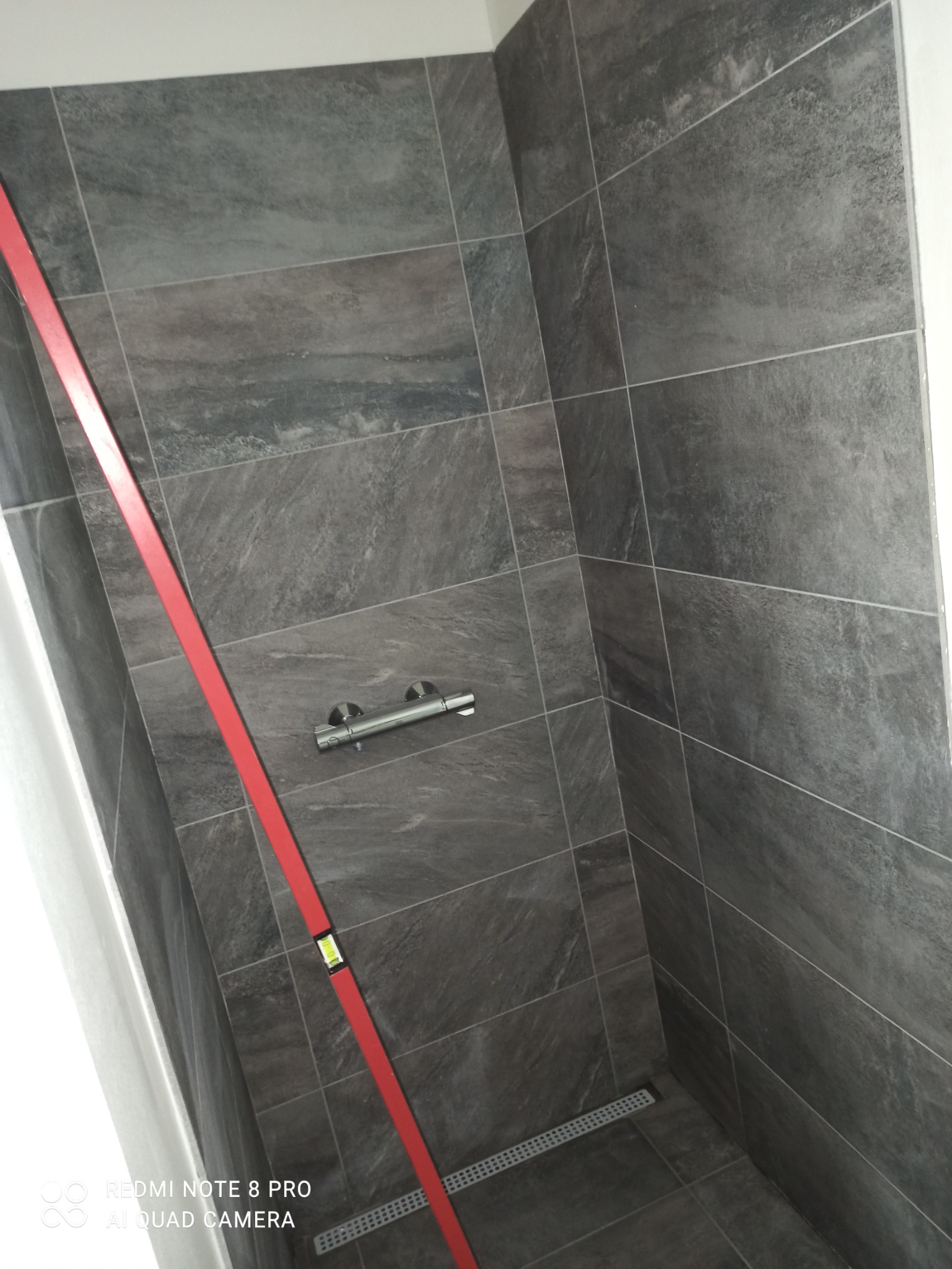 Ściana prysznica wyłożona szarymi płytkami imitującymi kamień, widoczna poziomica w kolorze czerwonym i odpływ liniowy w podłodze.