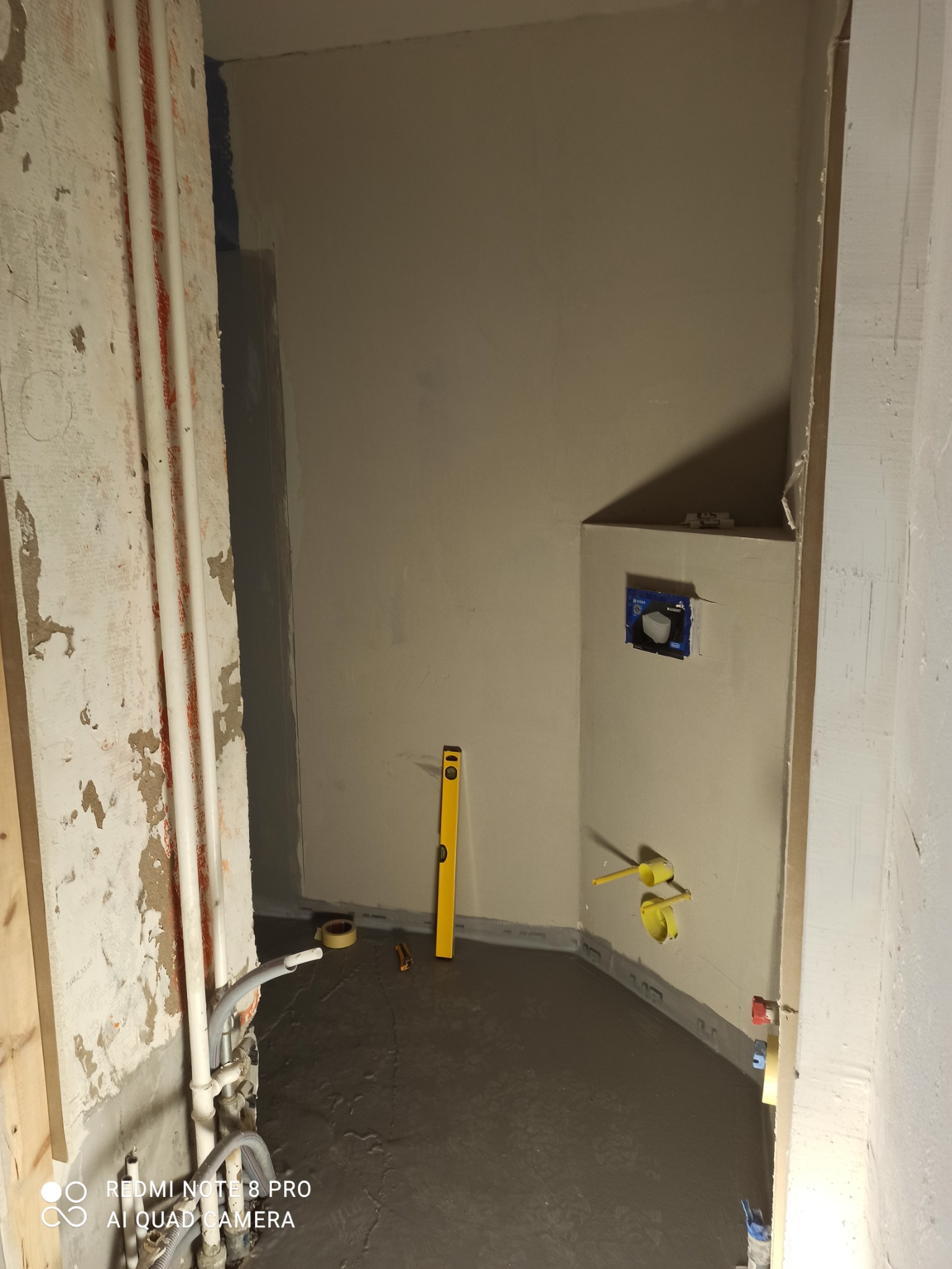 Wnętrze łazienki w trakcie remontu, z widocznymi rurami, wylewką podłogową, żółtą poziomicą opartą o ścianę i przygotowanym miejscem na podtynkowy stelaż WC.
