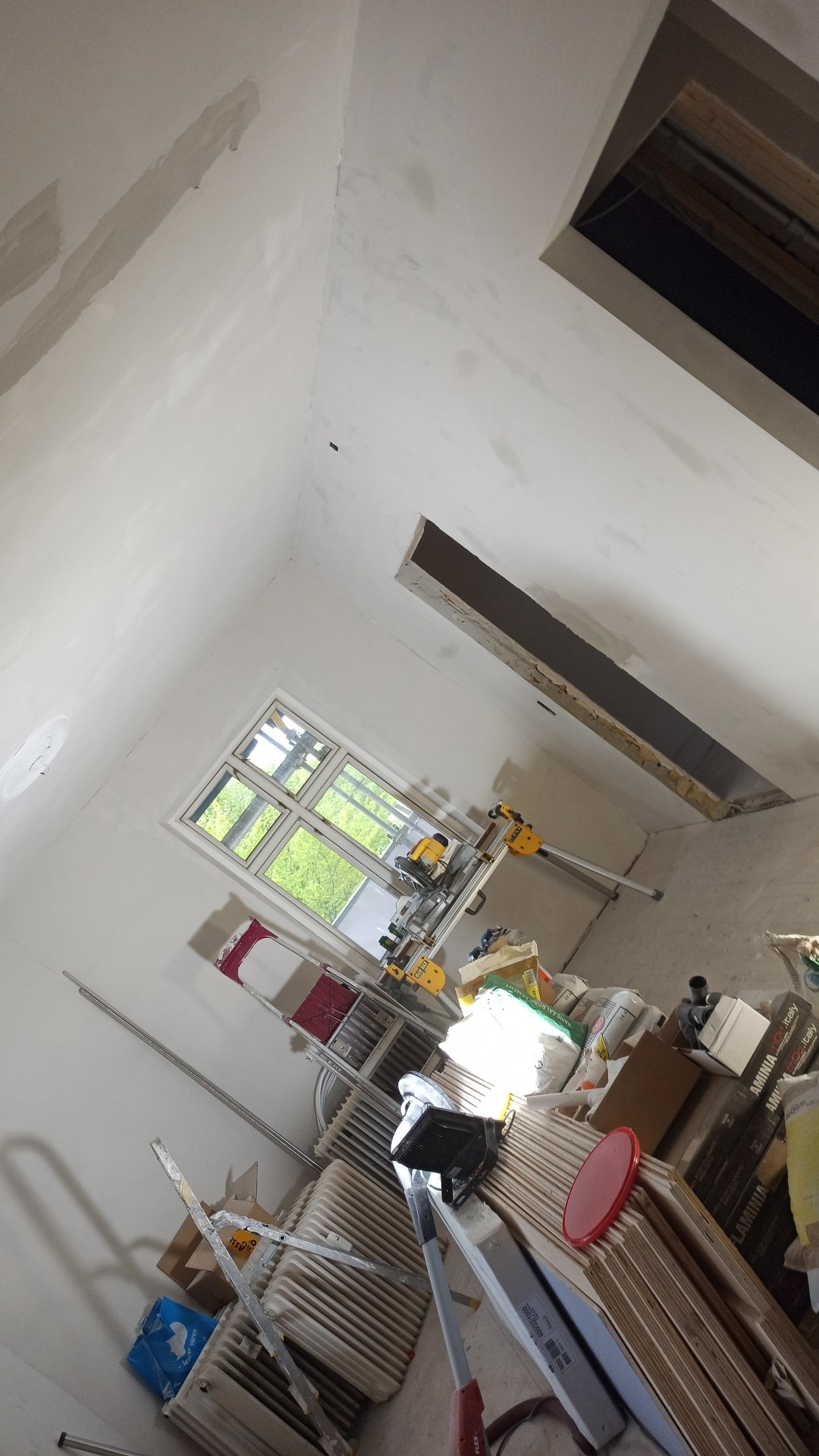 Wnętrze w trakcie remontu: szpachlowana ściana i sufit, okno z widokiem na zieleń, piła stołowa na stojaku, drabiny, grzejniki, materiały budowlane i otwór w suficie.