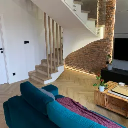 Nowoczesne, jasne schody z drewnianymi stopniami i szklaną balustradą, oświetlone od dołu, w salonie z ceglaną ścianą i parkietem w jodełkę.