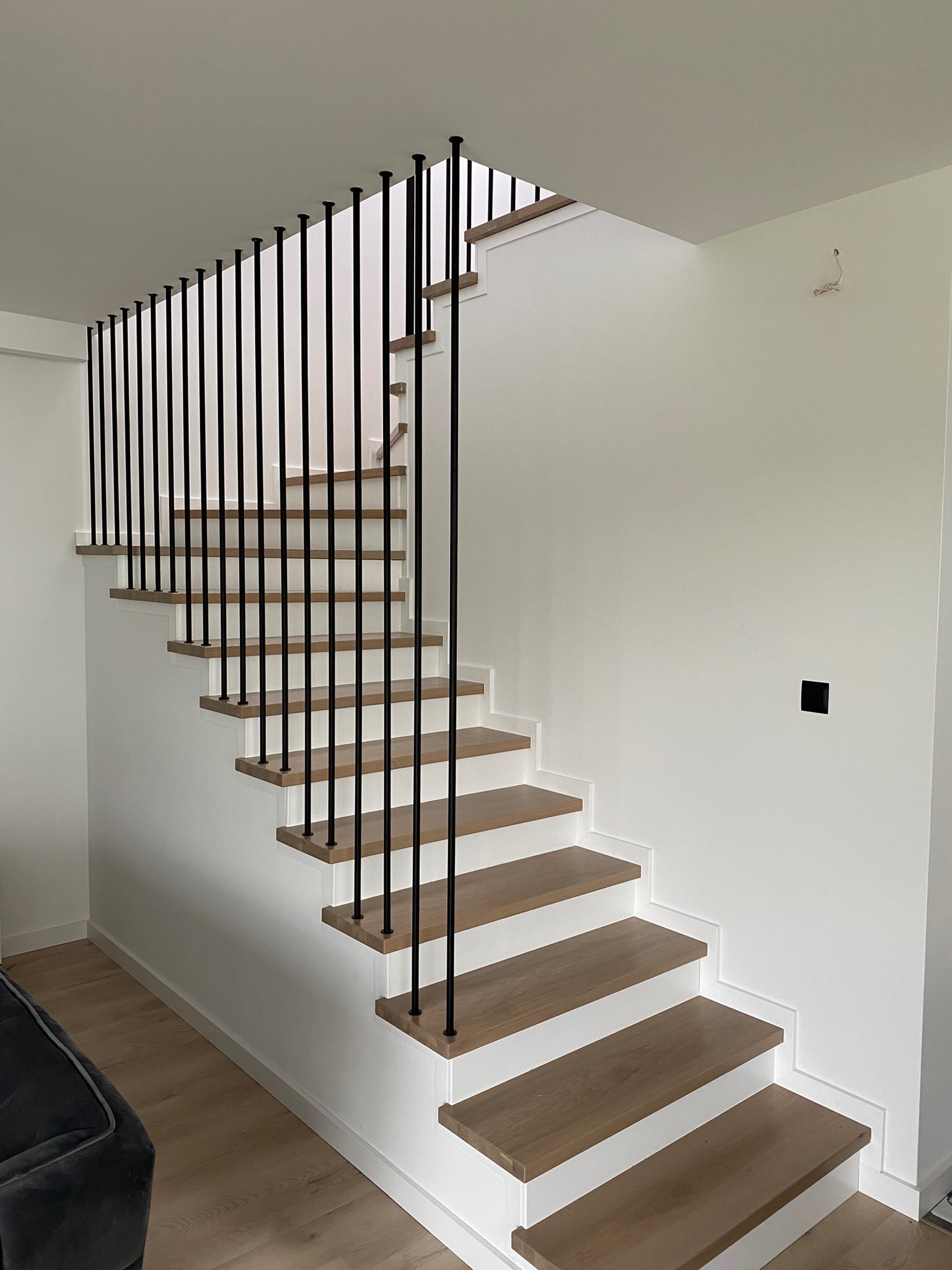 Nowoczesne, drewniane schody z czarnymi, metalowymi balustradami w minimalistycznym wnętrzu z białymi ścianami i drewnianą podłogą. Widoczne schody prowadzą na górę, a na ścianie wisi wystający kabel.